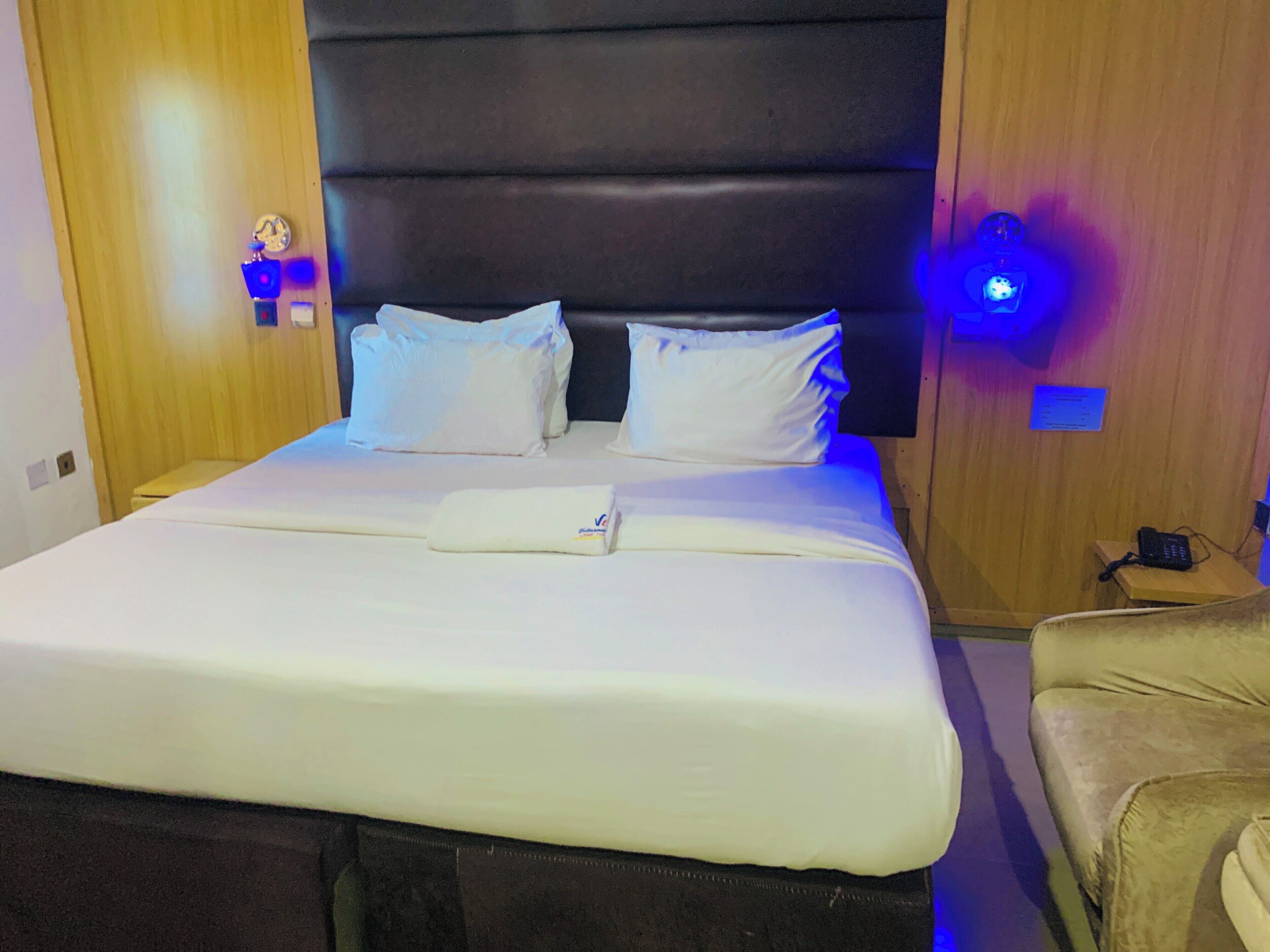 Una cama bien hecha con ropa blanca, un cabecero oscuro y una mesita de noche con un teléfono y una lámpara pequeña.