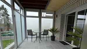 Une terrasse couverte avec un sol carrelé, deux tables accompagnées de chaises et des plantes en pot.