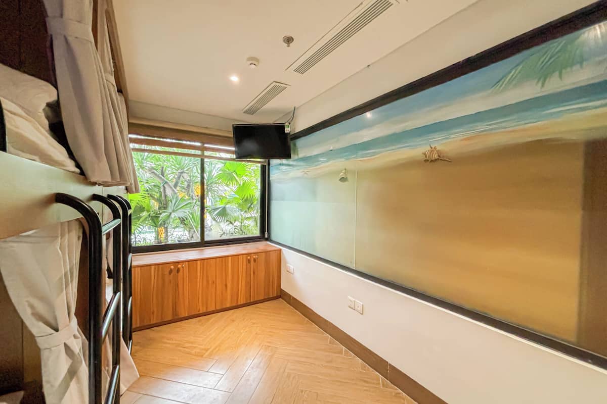 A hotel room with a large window, wooden flooring, a built-in bench, and a wall-mounted TV.