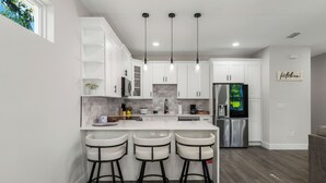 Una cocina moderna con armarios blancos, una isla grande con taburetes de bar, electrodomésticos de acero inoxidable e iluminación colgante.