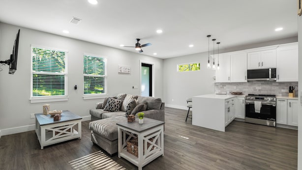 Una sala moderna con un sofá gris, una mesa de centro blanca y una mesita auxiliar. Hay una zona de cocina con armarios blancos, un microondas y una estufa.