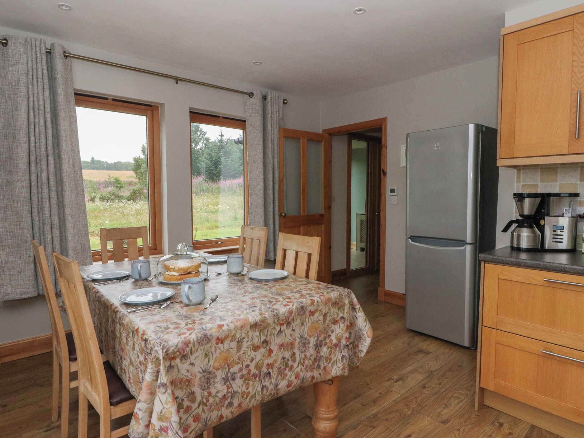 A dining area with a table set for two, a refrigerator, wooden cabinets, and a view of a garden through the windows.