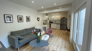 Un salon moderne avec un canapé gris, une table basse ronde et un fauteuil rose. On y trouve également un coin cuisine avec des meubles blancs et un réfrigérateur en acier inoxydable.