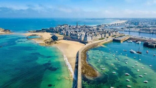 Vista aérea de una ciudad costera con una fortificación destacada, una playa de arena y un puerto deportivo con numerosos barcos.