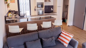 Una cocina moderna con una isla central, taburetes blancos y un sofá gris con un cojín a rayas naranjas.