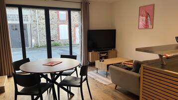 Un salon moderne avec une table ronde, des chaises, un canapé, un téléviseur et une vue sur une rue bordée d’immeubles.