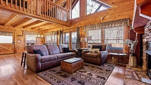 A log cabin interior with a wooden ceiling, a large window with blinds, a leather sofa, a small table, and a fireplace. - Southern Memories – 2BR Cabin, Resort Pool, King Beds, Game Room (Pigeon Forge)