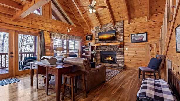 A cozy cabin interior with a stone fireplace, wooden walls, a dining area, and a large window overlooking a snowy landscape. - Southern Memories – 2BR Cabin, Resort Pool, King Beds, Game Room (Pigeon Forge)