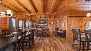 A cozy wooden cabin interior with a dining area, a fireplace, and a stone wall with a sunset photo. - Southern Memories – 2BR Cabin, Resort Pool, King Beds, Game Room (Pigeon Forge)