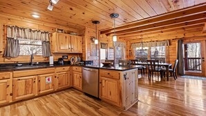 A cabin-style kitchen with wooden cabinets, a central island, and a dining area with a table and chairs. - Southern Memories – 2BR Cabin, Resort Pool, King Beds, Game Room (Pigeon Forge)