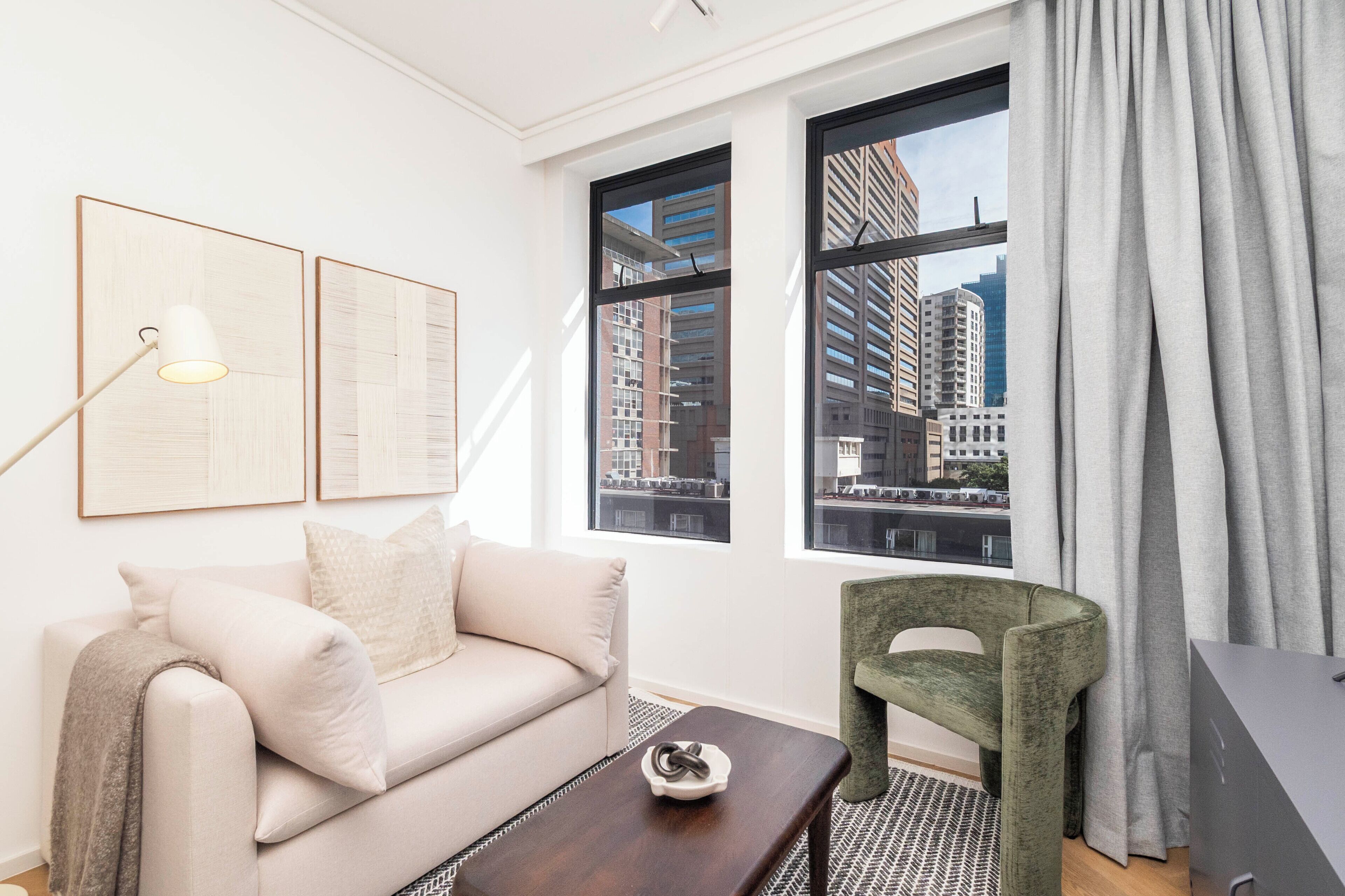 A modern living room with a sofa, a small table, a green chair, and a large window with a city view.