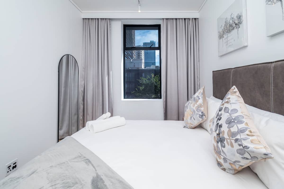 Een moderne hotelkamer met een groot bed, grijze gordijnen, uitzicht op gebouwen door een raam en een flatscreen televisie.