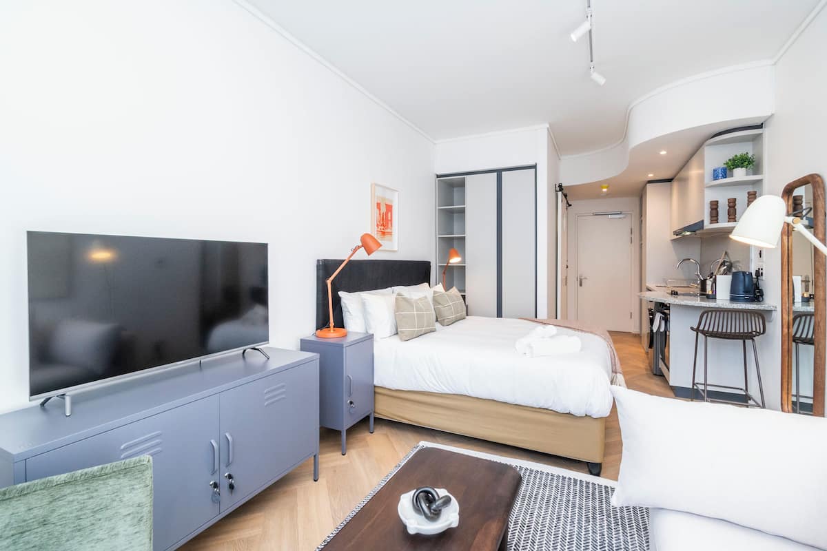 Een moderne hotelkamer met een grote flatscreen televisie, een bed met wit beddengoed, een grijze kast en een kleine kitchenette.