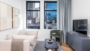 Ein modernes Wohnzimmer mit einer Couch, einem grünen Sessel, einem kleinen Tisch und einem Fernseher auf einer Kommode. Große Fenster bieten Blick auf Gebäude und eine Stadtansicht.