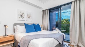 Ein modernes Schlafzimmer mit einem großen Bett, einem Nachttisch, einer Lampe und Blick auf eine Stadtlandschaft durch ein großes Fenster.