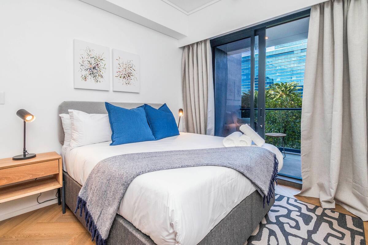 Een moderne slaapkamer met een groot bed, een nachtkastje, een lamp en een stadsgezicht door een groot raam.