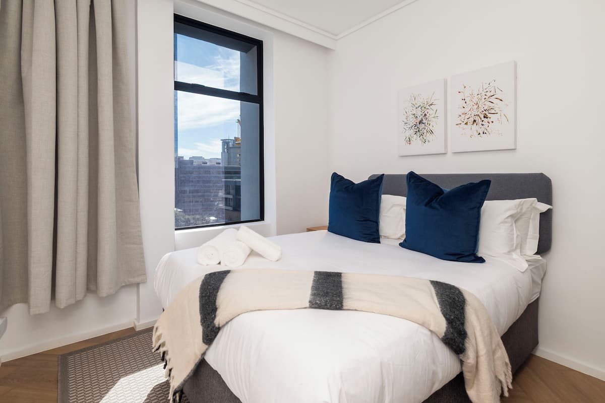 Een moderne hotelkamer met een groot bed, uitzicht op de stad door een raam, en twee kunstwerken aan de muur.