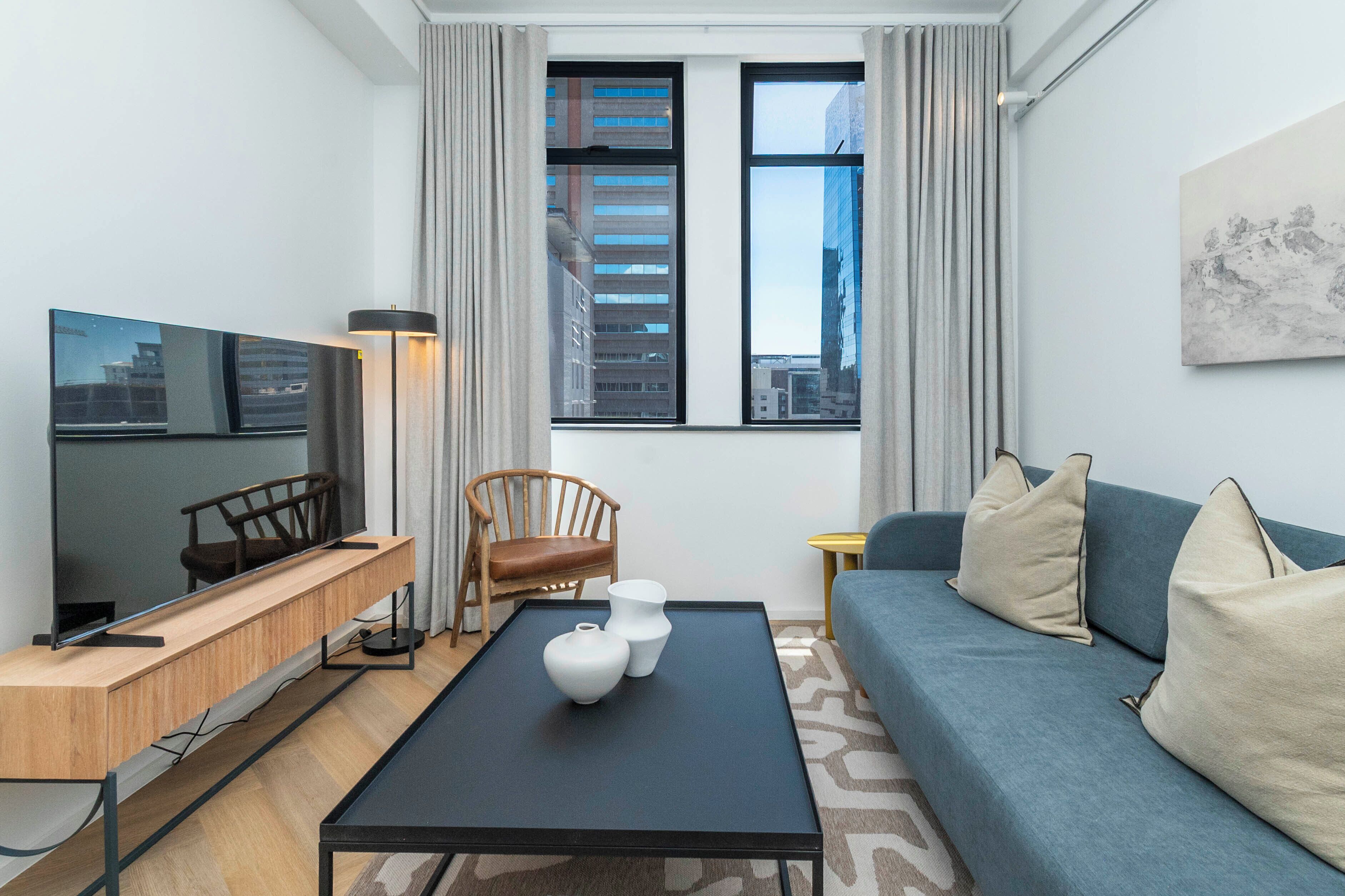 A modern living room with a flat-screen TV, a wooden coffee table, a blue sofa, and a city view through large windows.