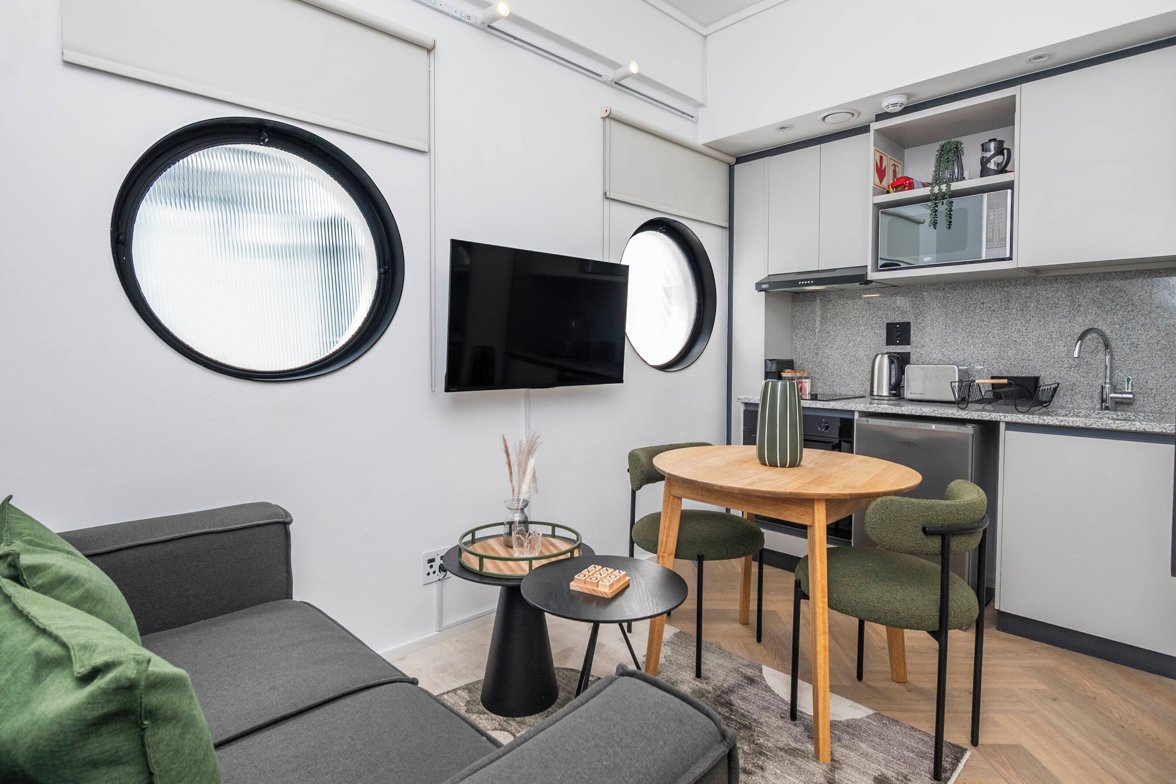 A compact living space with a sofa, a round dining table with chairs, a TV mounted on the wall, and a kitchenette with a microwave and sink.