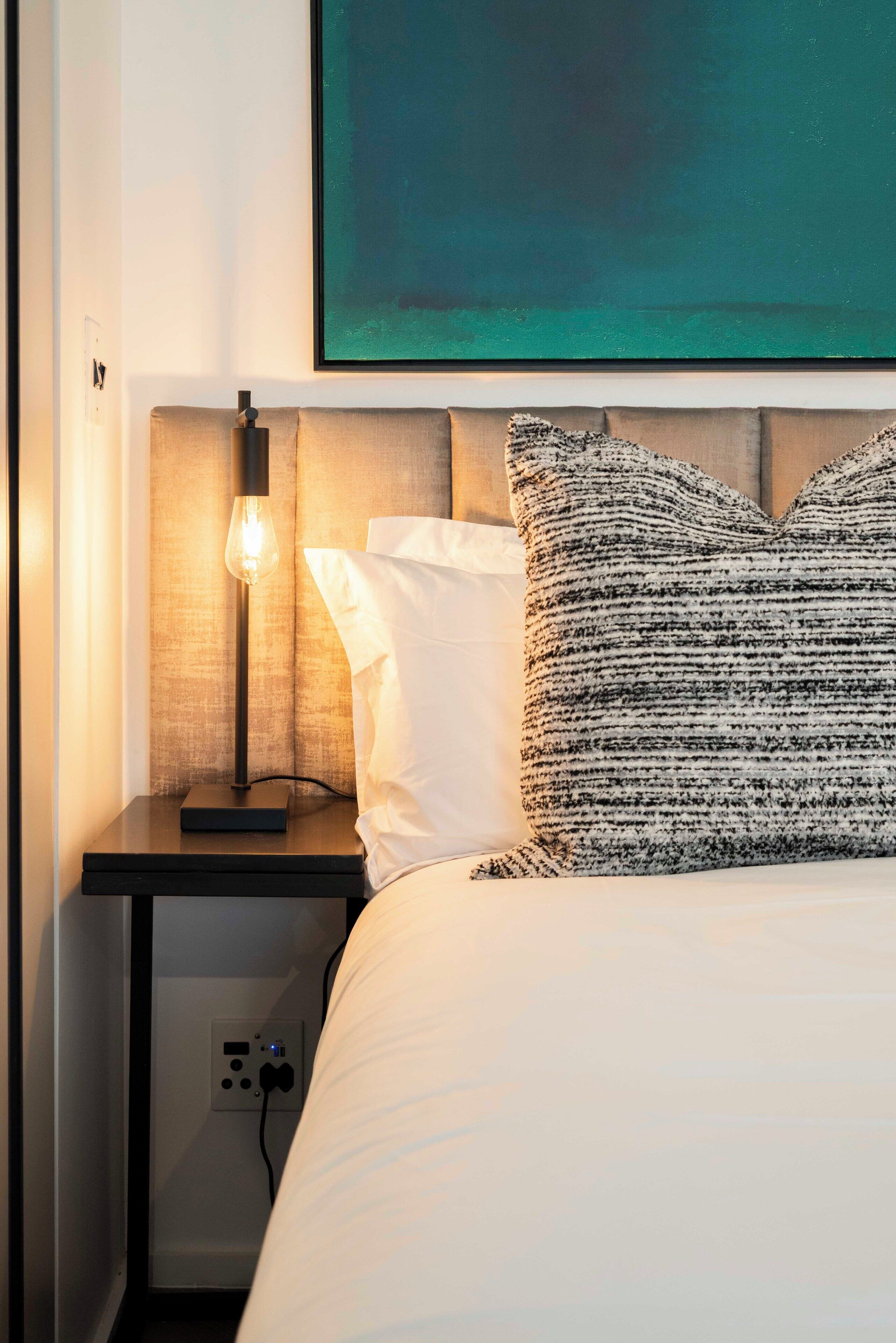 A neatly made bed with a textured pillow, a bedside lamp, and a power outlet on a nightstand.
