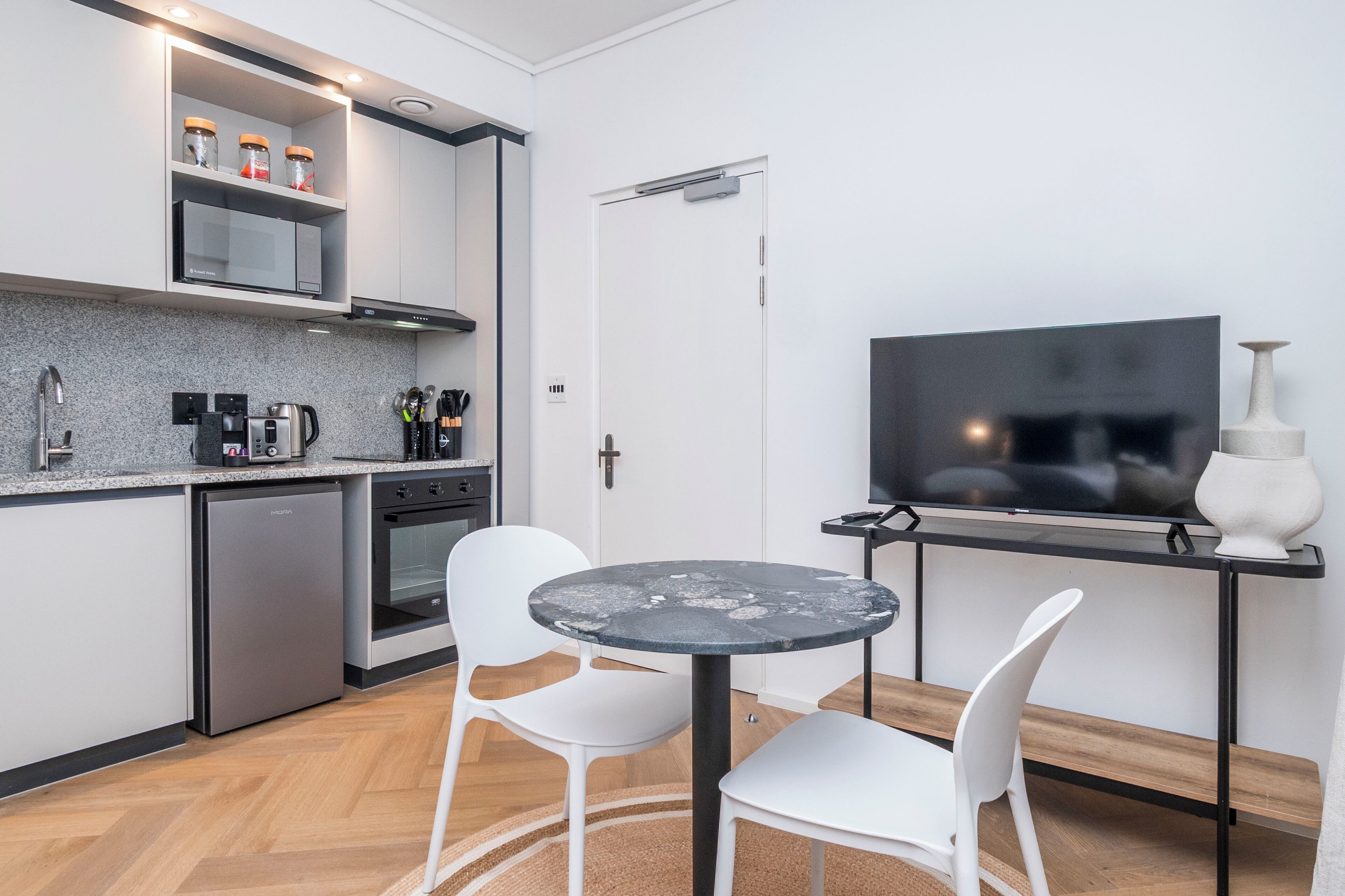 A modern kitchen with a microwave, coffee maker, and cabinets. There is a dining table with chairs and a TV on a stand.