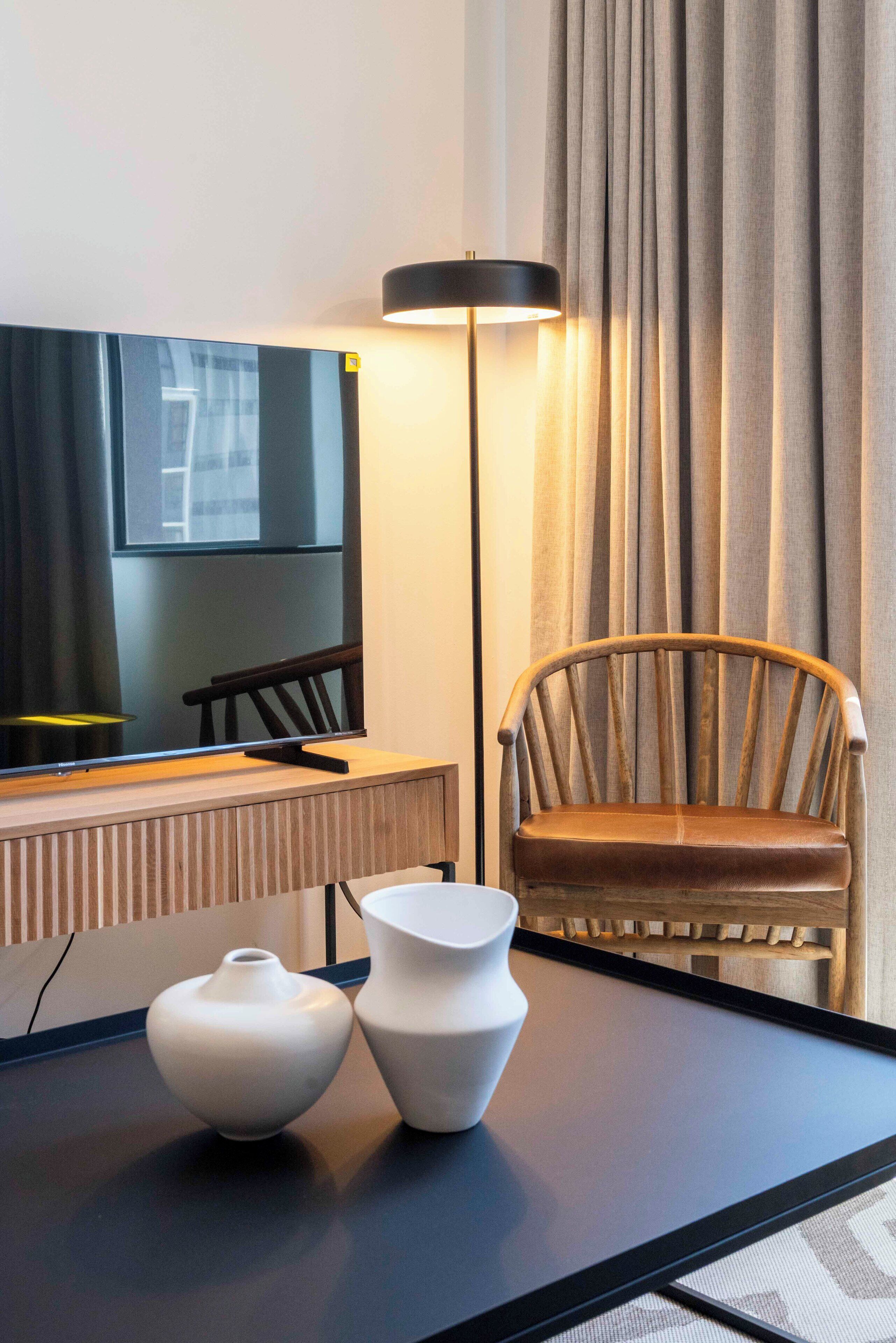 Ein modernes Hotelzimmer mit einem großen Flachbildfernseher, einem Holzstuhl und zwei weißen Vasen auf einem dunklen Tisch.