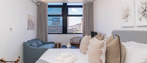 A modern hotel room with a large bed, a blue sofa, a small table, and a view of the city through large windows.