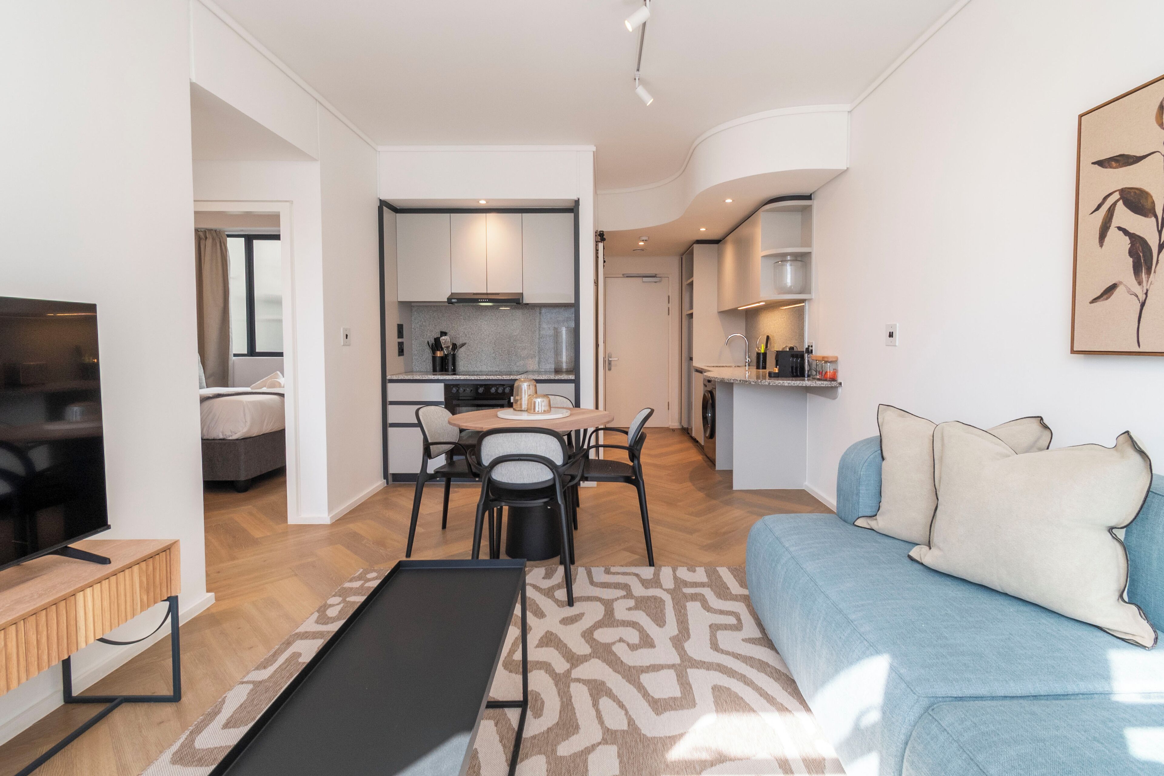 Una sala moderna con sofá, una mesa de centro y televisor. Hay un comedor con mesa y sillas. Se ve la cocina al fondo, con armarios y estufa.