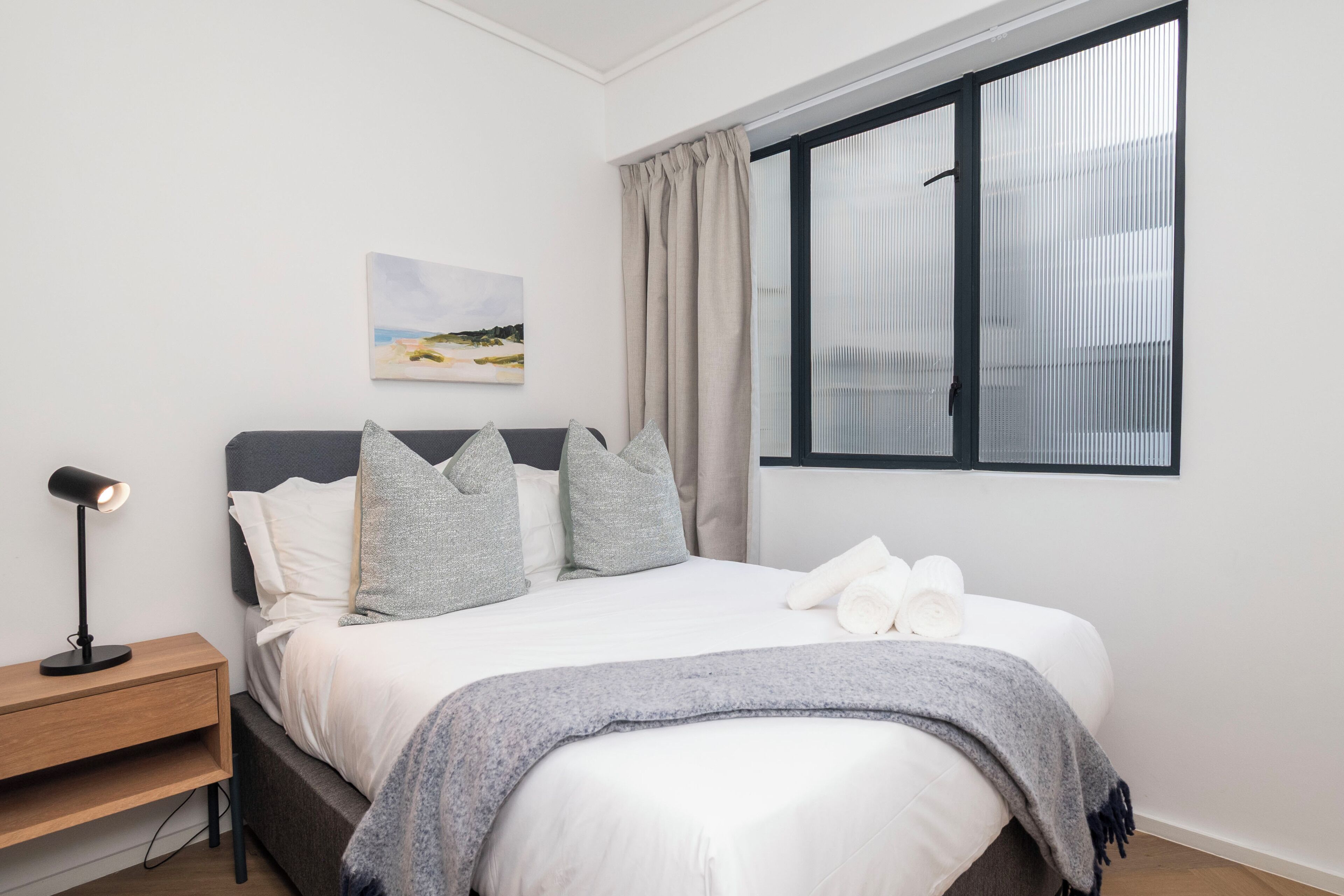 A neatly made bed with pillows and a blanket, a bedside lamp, and a window with curtains.
