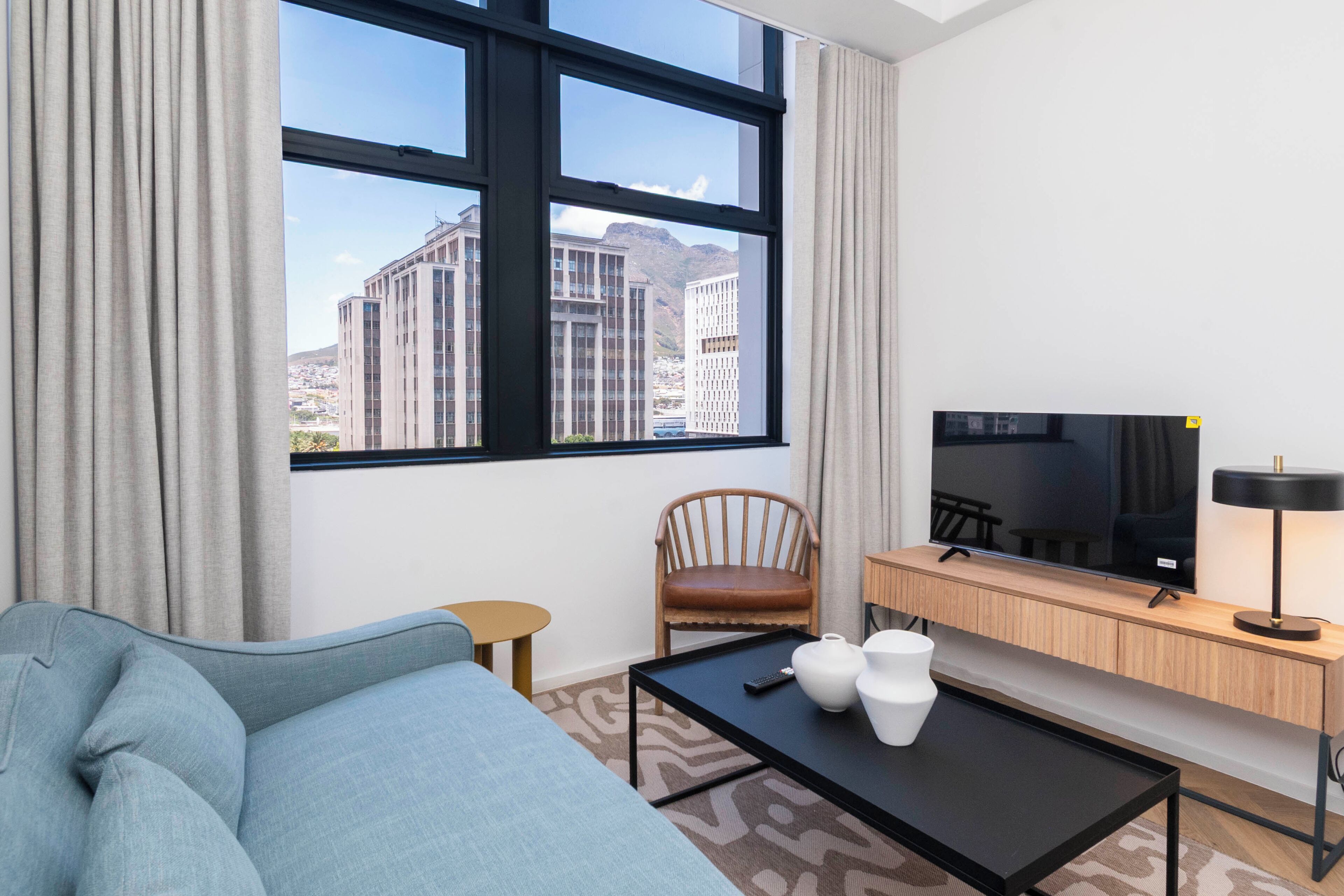 Habitación de hotel con un ventanal grande, un sofá, una mesita, un televisor y una lámpara.