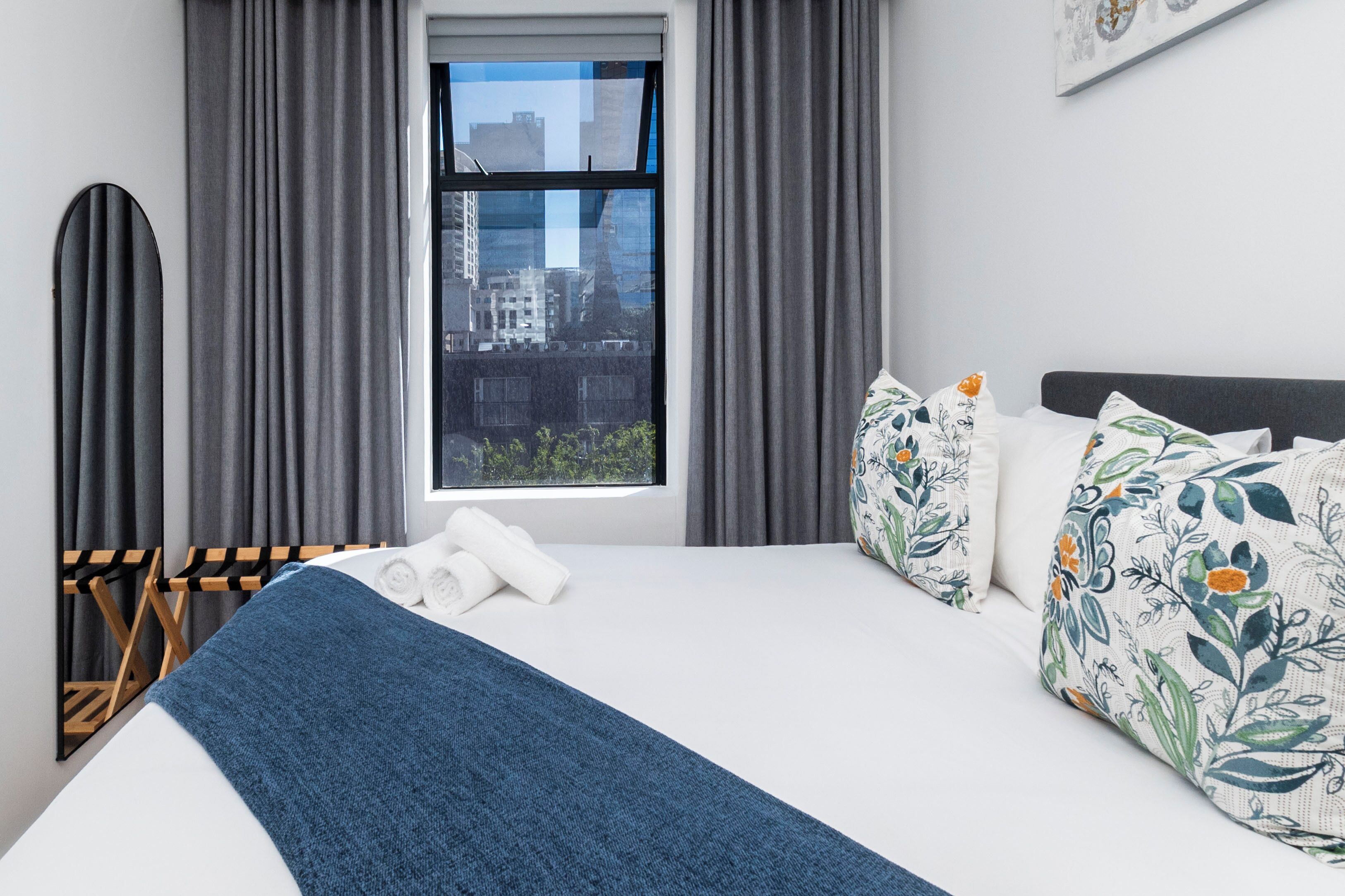Een hotelkamer met een groot bed, grijze gordijnen, een raam met uitzicht op de stad en een blauwe deken.