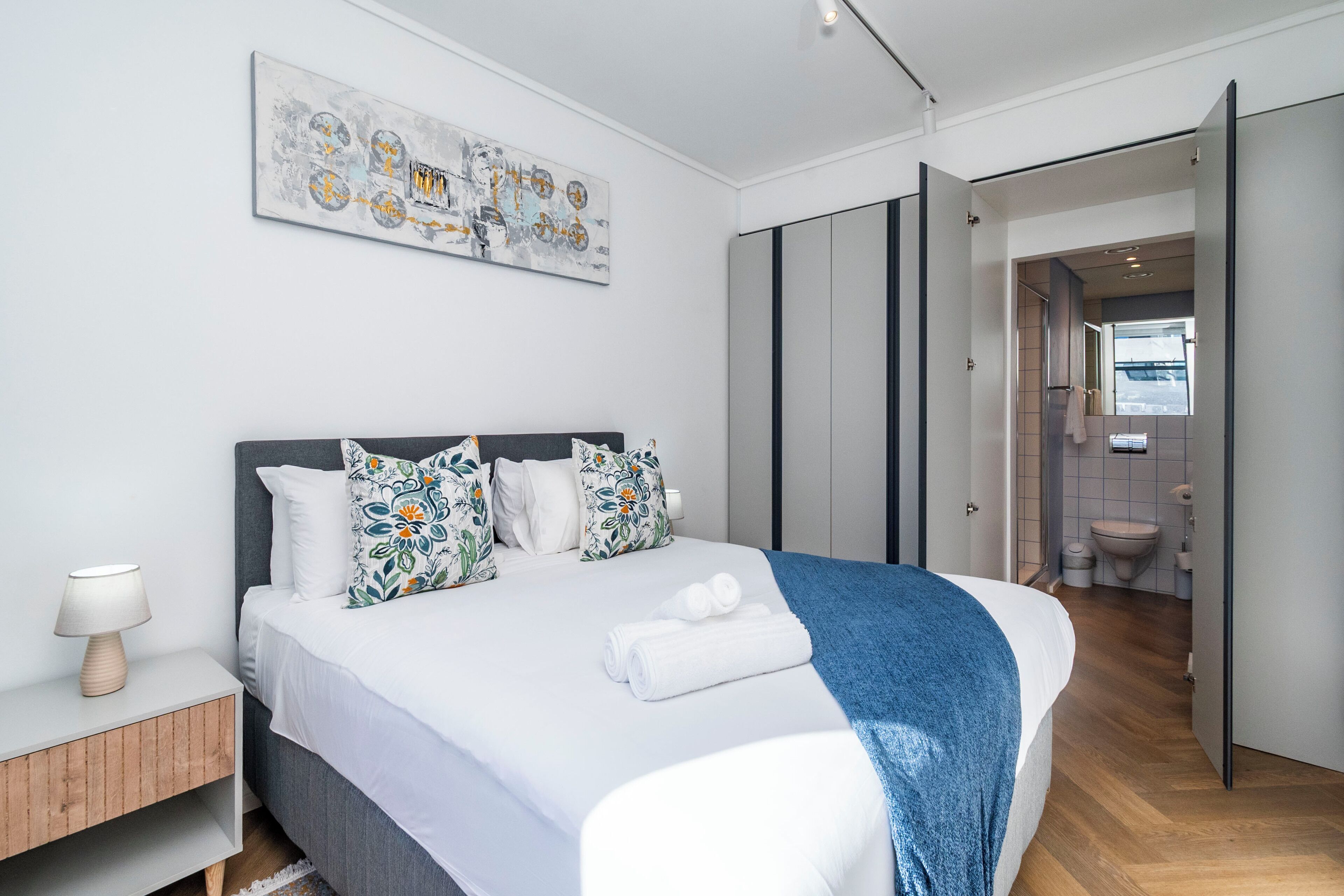 Een moderne slaapkamer met een groot bed, een nachtkastje met een lamp, en een badkamer die door een open deur zichtbaar is.