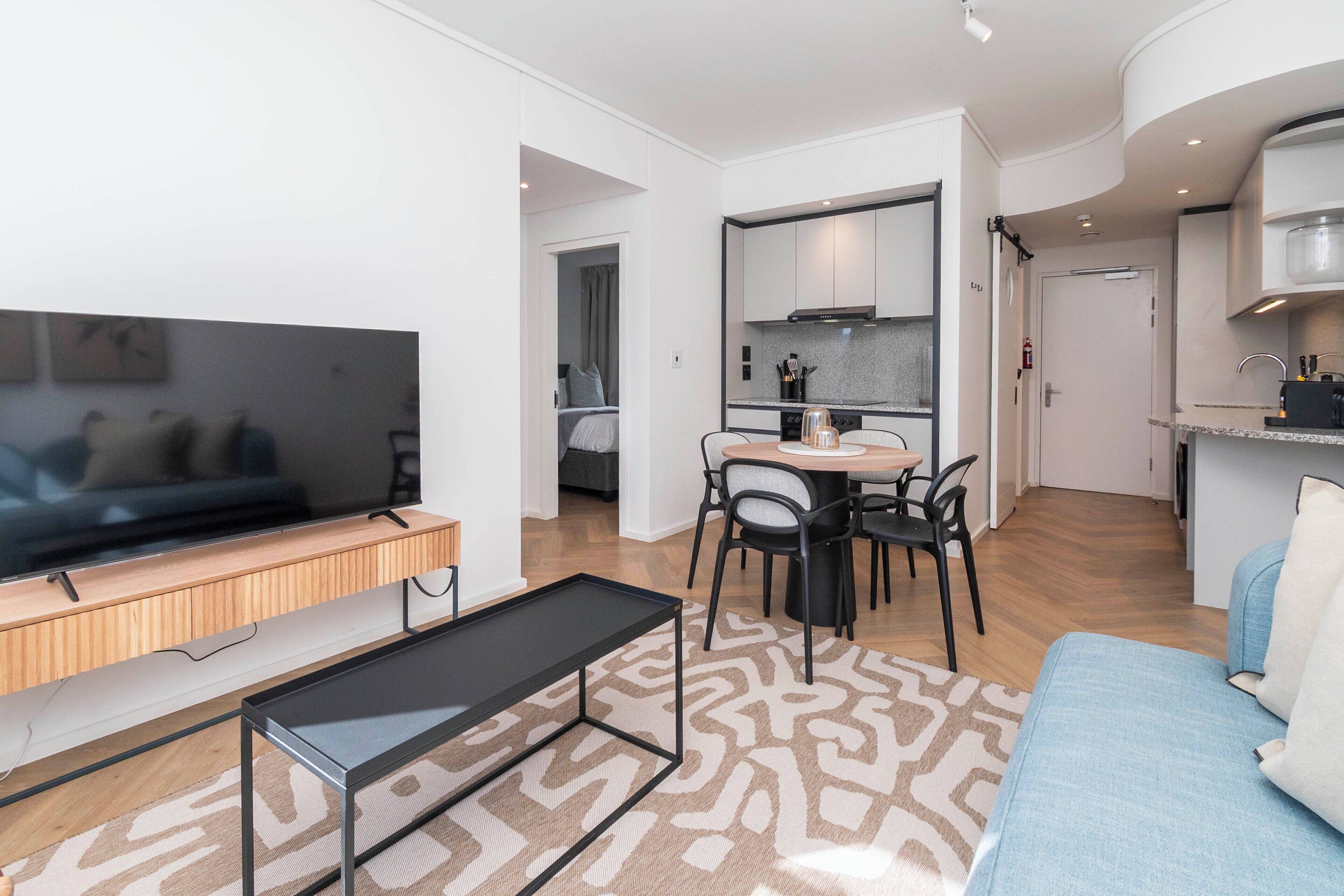 Een moderne woonkamer met een flatscreen televisie, een bank, een eettafel met stoelen en een keukenhoek met kasten en een aanrecht.