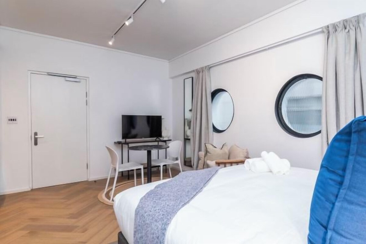 A modern hotel room with a bed, a desk with a chair, a television, and a round mirror.