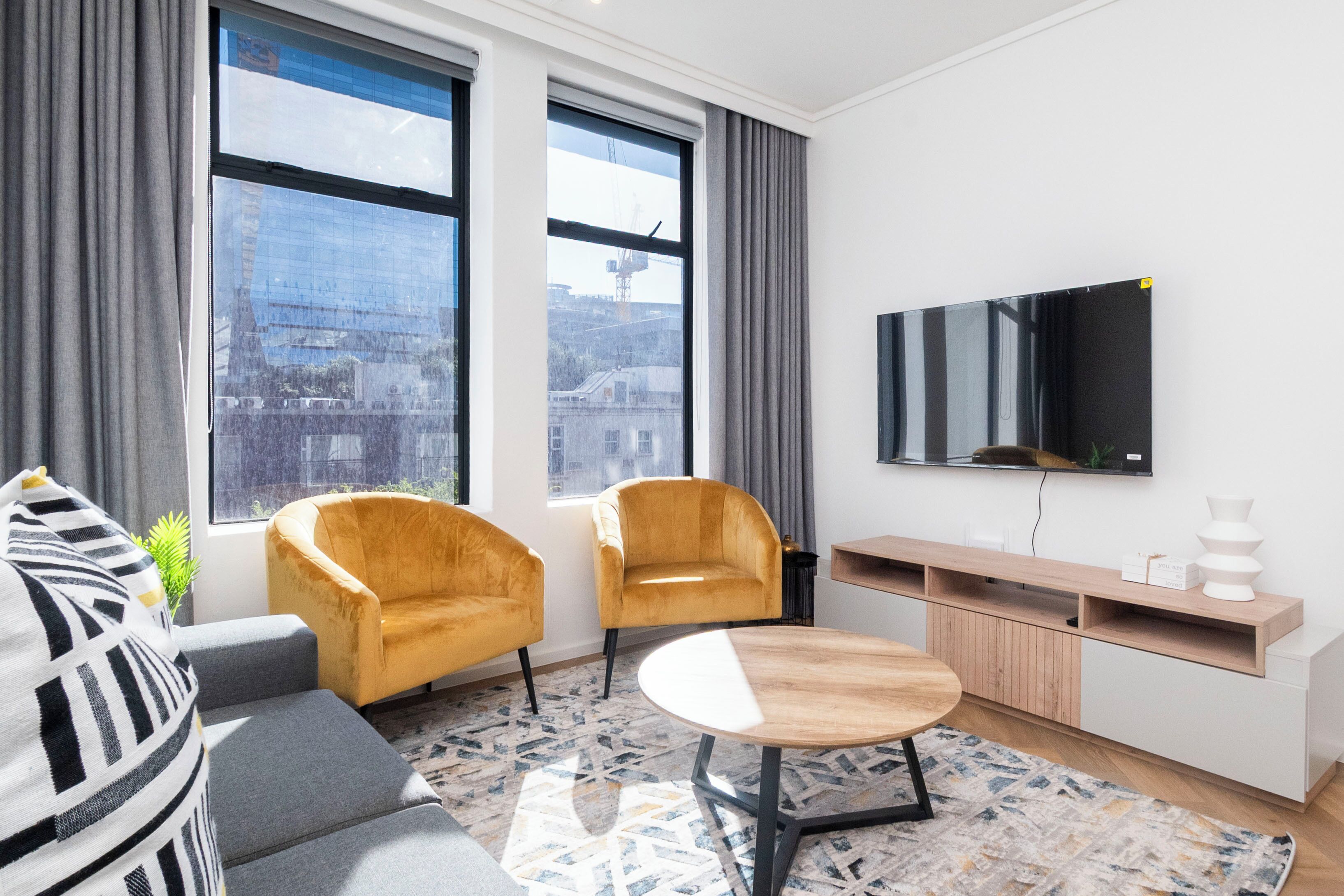 A modern living room with a sofa, two armchairs, a coffee table, a TV, and a city view through large windows.