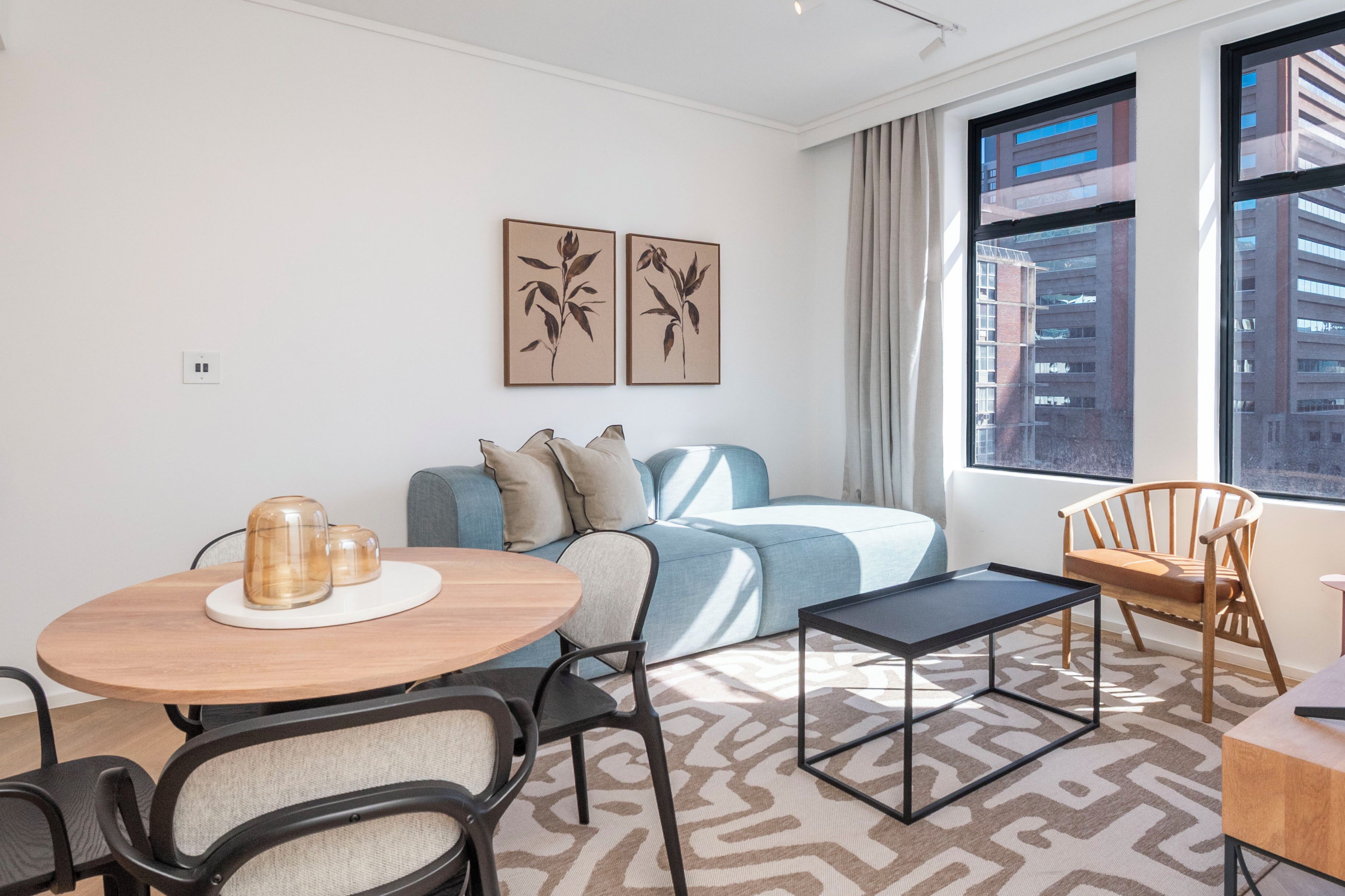 A modern living room with a sofa, a coffee table, and a dining area with chairs. There are two framed artworks on the wall and a large window with a view of buildings.