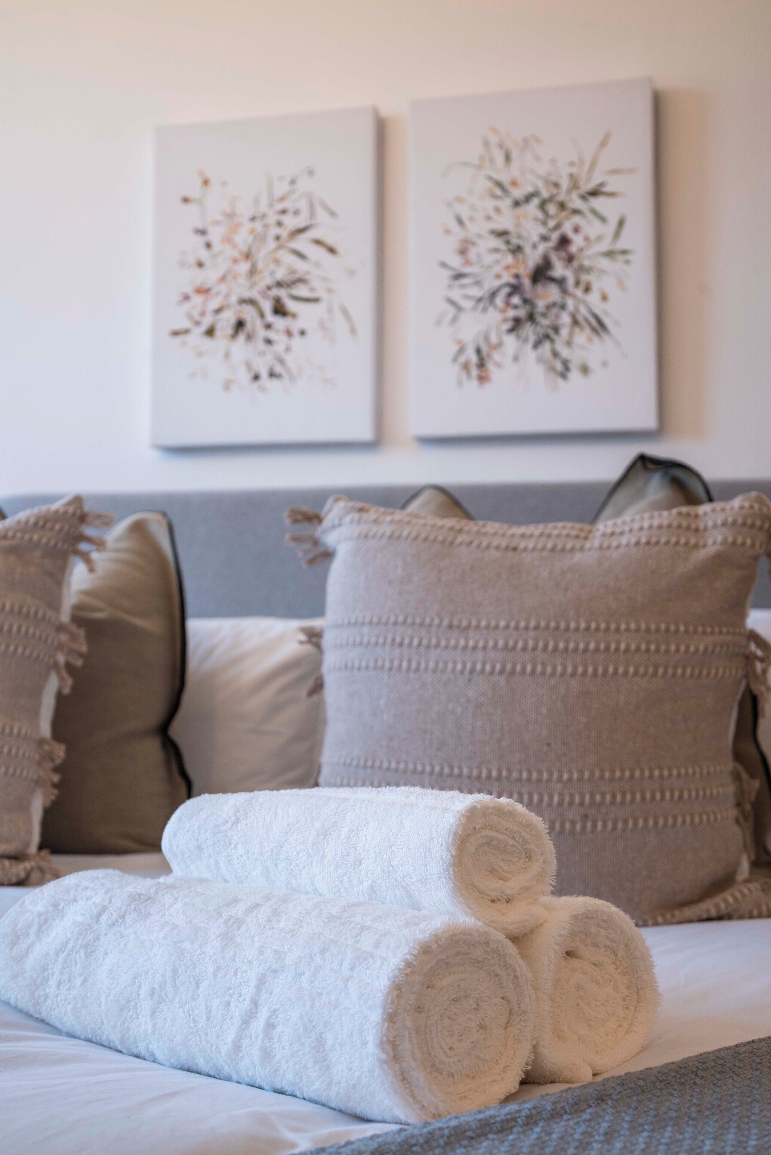 A neatly made bed with rolled white towels, a grey sofa with patterned cushions, and two abstract floral wall art pieces.