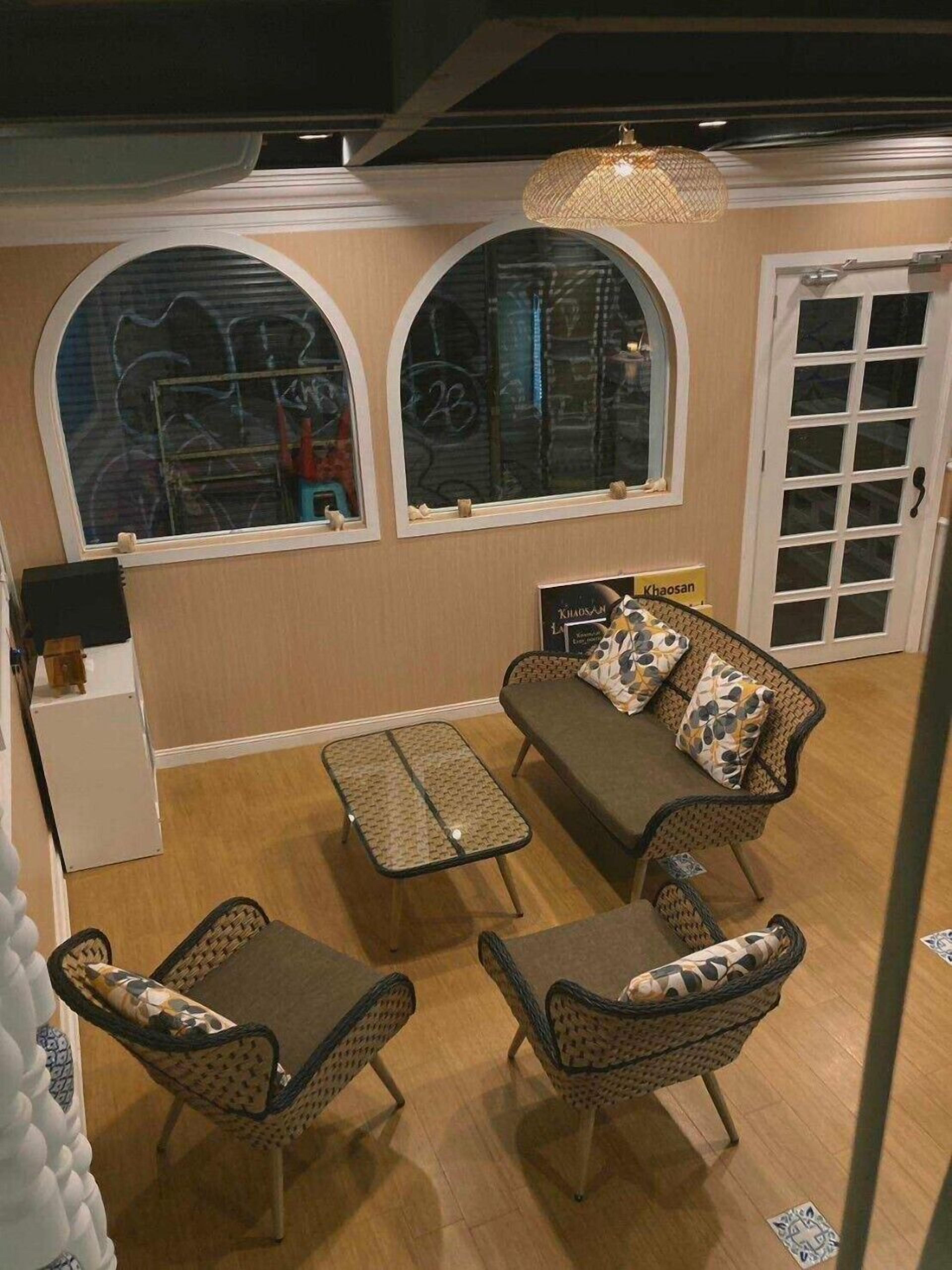 Une pièce chaleureuse avec un canapé, deux fauteuils, une petite table et une lampe décorative. On y trouve deux fenêtres en arc surmontées de graffitis, une porte et une petite armoire blanche.