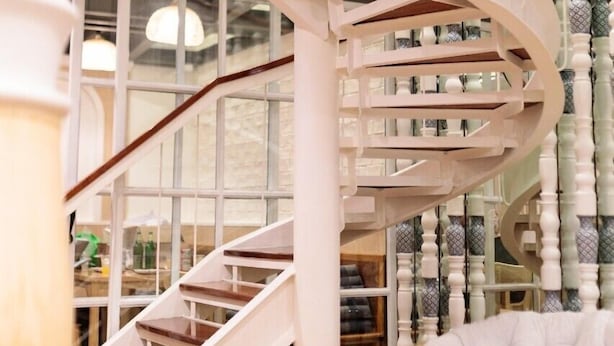 A spiral staircase with white railings and wooden steps, leading to a room with large windows and a wicker chair. - ReRe Hostel (Bangkok)