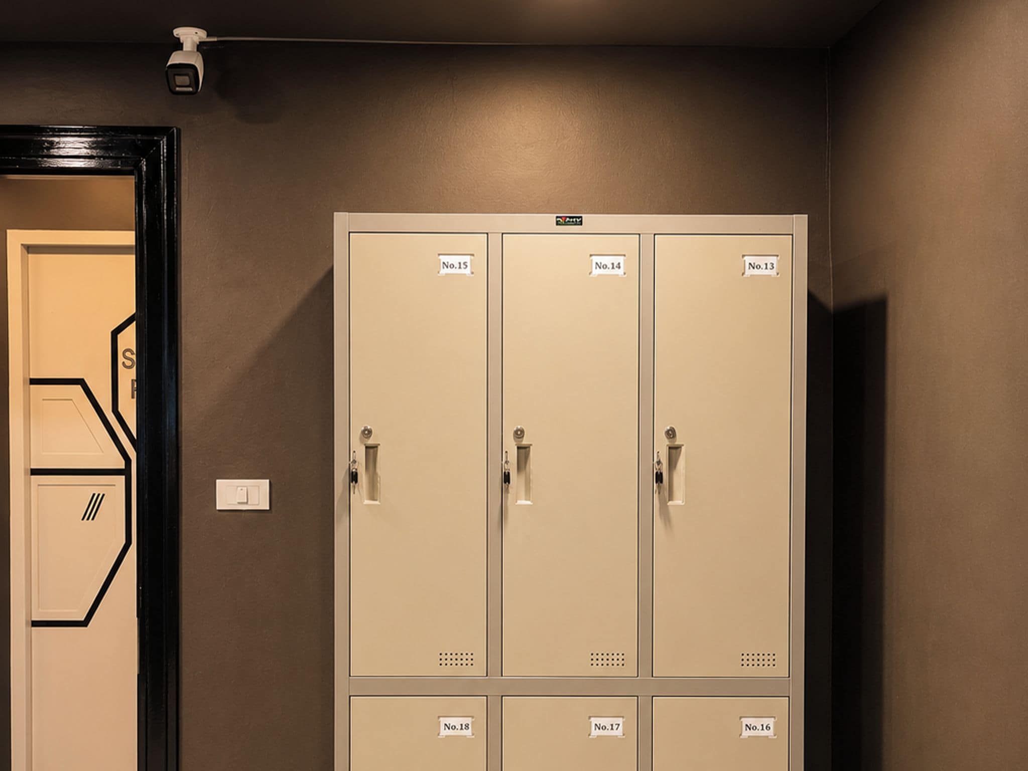Una serie di armadietti beige con etichette numerate, una porta con un motivo geometrico e un interruttore della luce sulla parete.