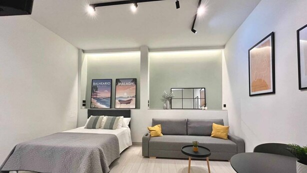 A modern hotel room with a bed, a sofa, a small table, and a chair. There are posters on the wall and a mirror above the sofa. - Mena Trip Malaga- Studio in the historic center (Málaga)