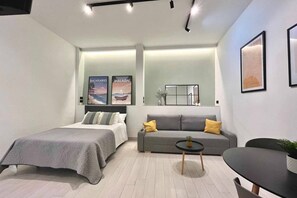 A modern hotel room with a bed, a sofa, a small table, and a chair. There are posters on the wall and a mirror above the sofa.