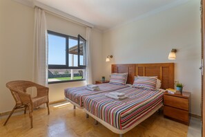 Un dormitorio con cama, una silla, una mesita de noche y una lámpara, además de un ventanal con cortinas.