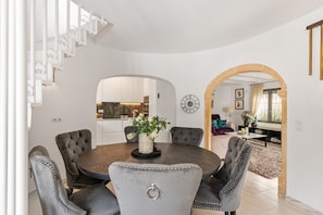 A dining area with a round table, six upholstered chairs, a vase with flowers, and a clock on the wall.