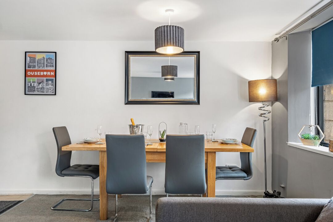 A dining area with a wooden table, four chairs, a hanging light fixture, a framed picture on the wall, and a floor lamp.