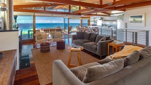 Amplia sala de estar con un gran ventanal con vista al océano, un rincón de descanso acogedor y una cocina moderna al fondo.