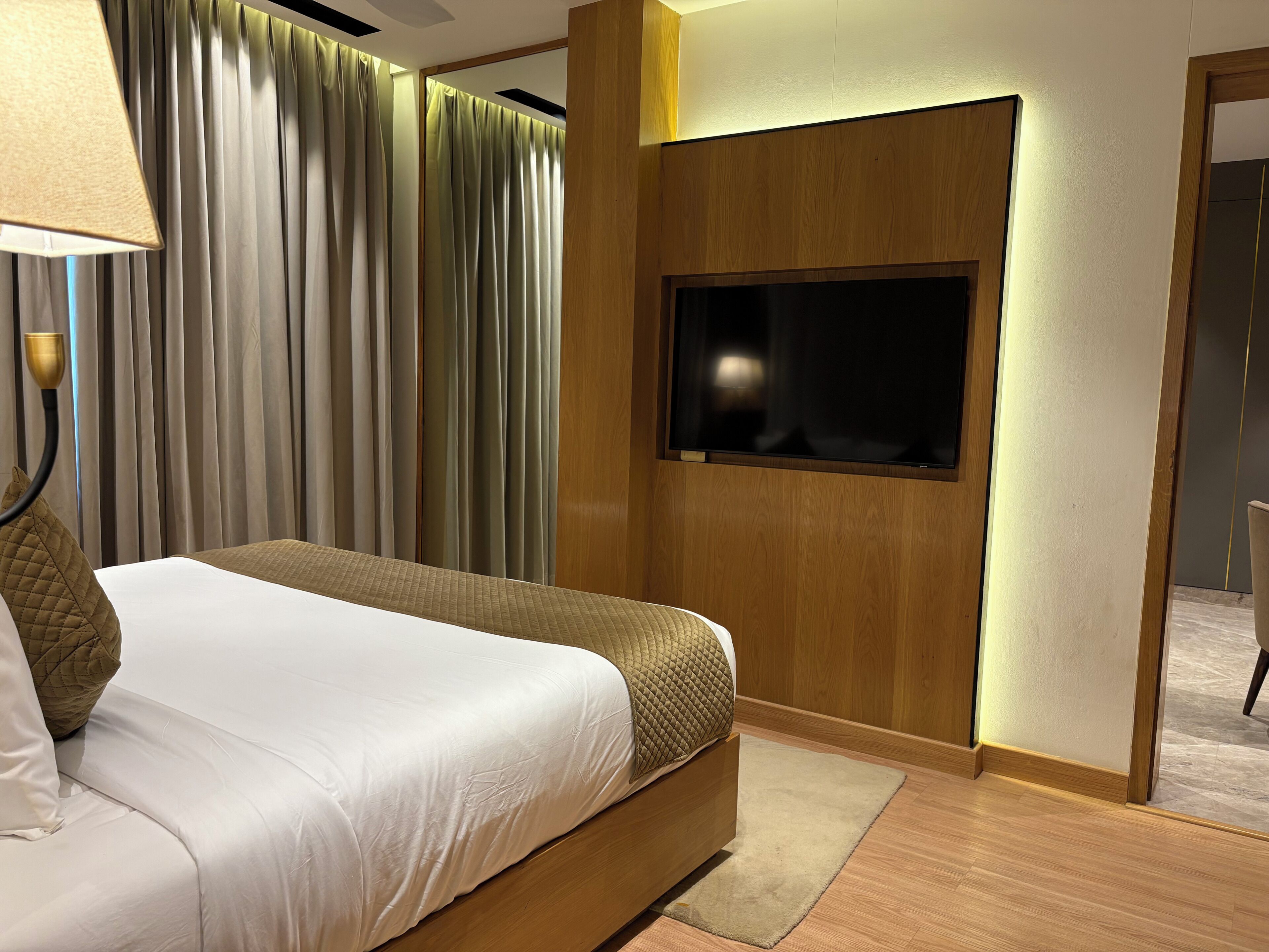 Habitación de hotel con cama, cortinas, un televisor montado en un panel de madera y una lámpara.