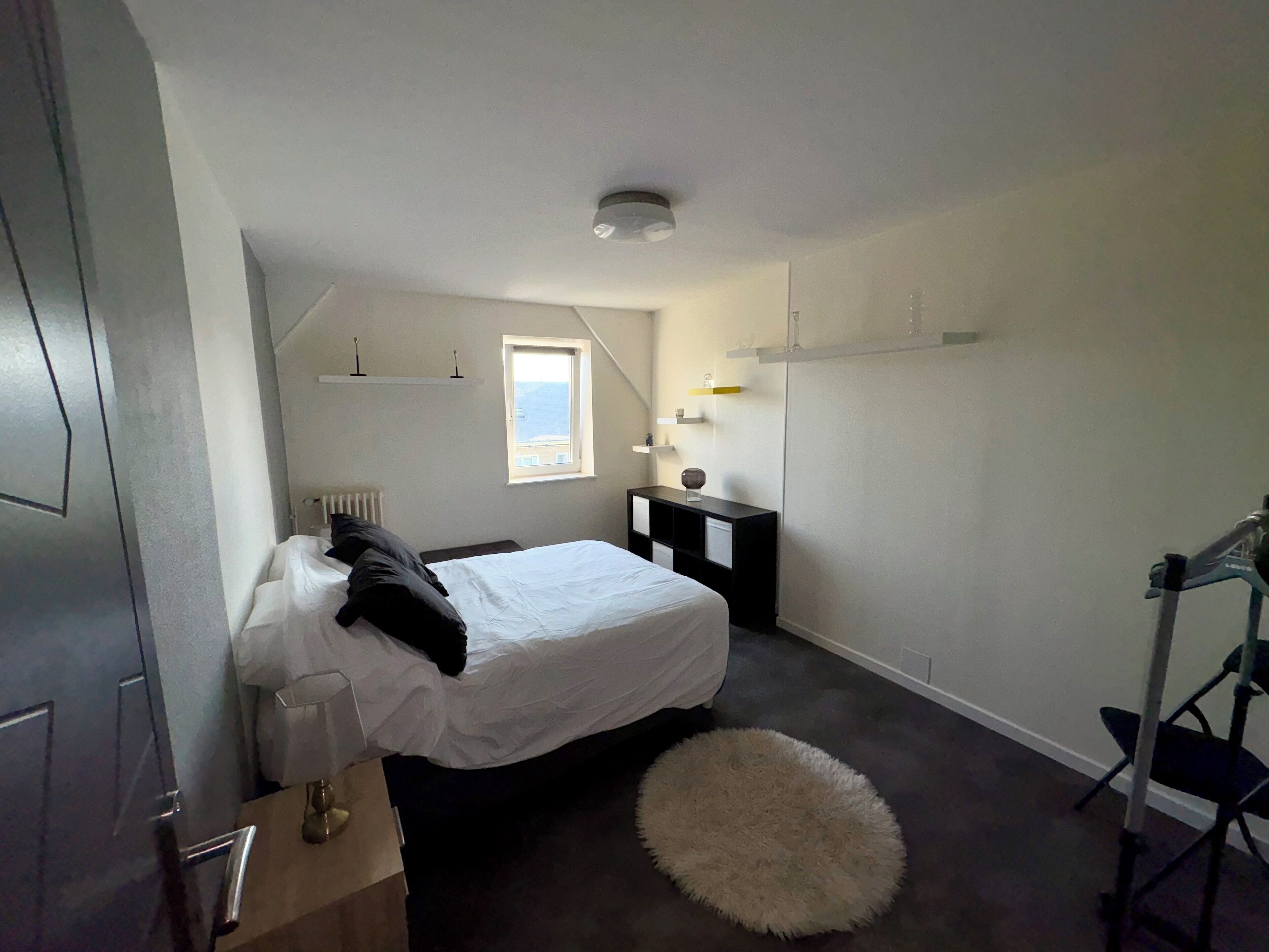 Une chambre à coucher comprenant un lit, un petit bureau, une chaise, un radiateur, une fenêtre et un luminaire au plafond.