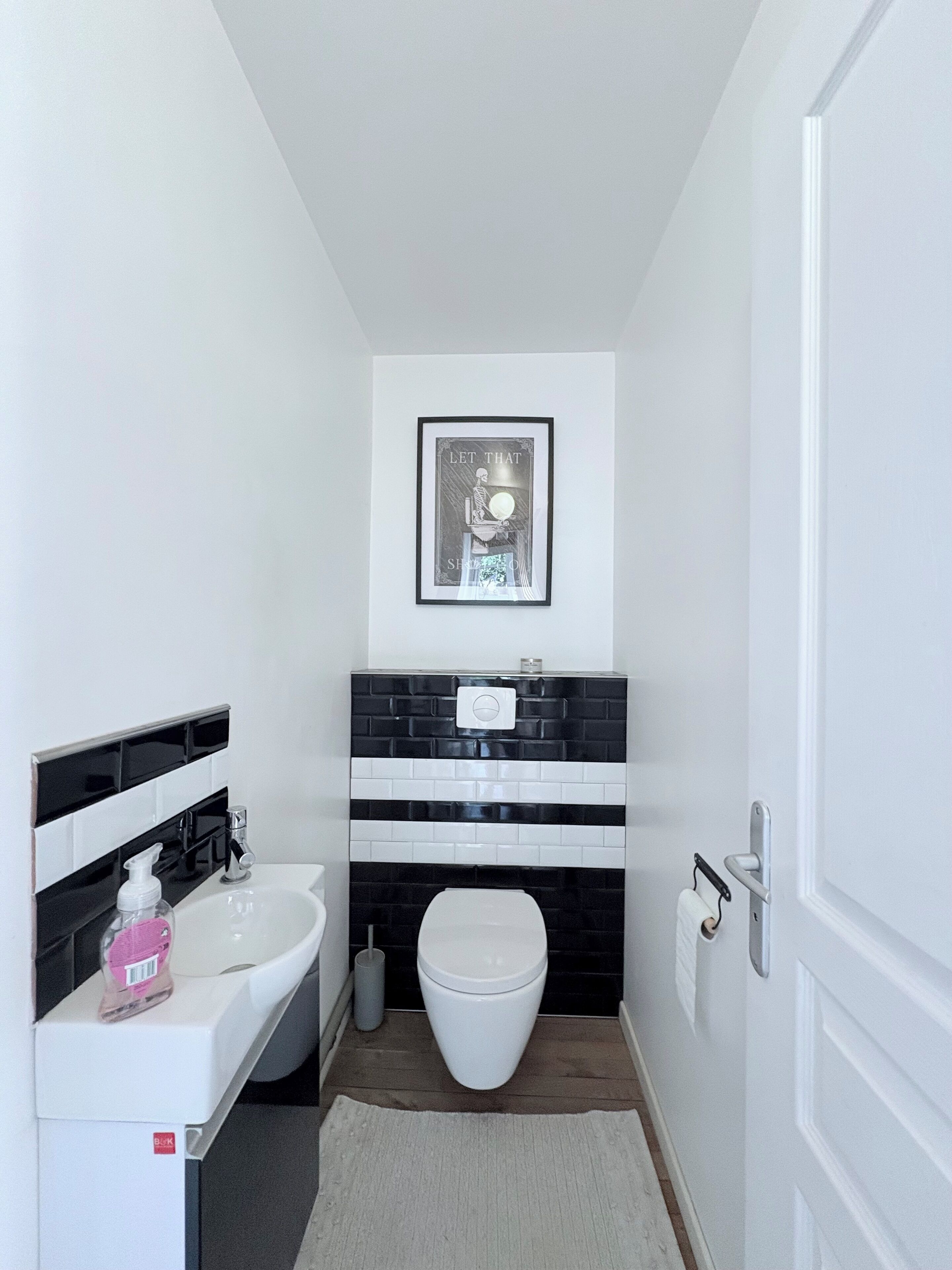 Une petite salle de bain avec un lavabo blanc, un pare-chocs en carreaux noirs et blancs, une œuvre d’art encadrée au-dessus des toilettes et une porte blanche.