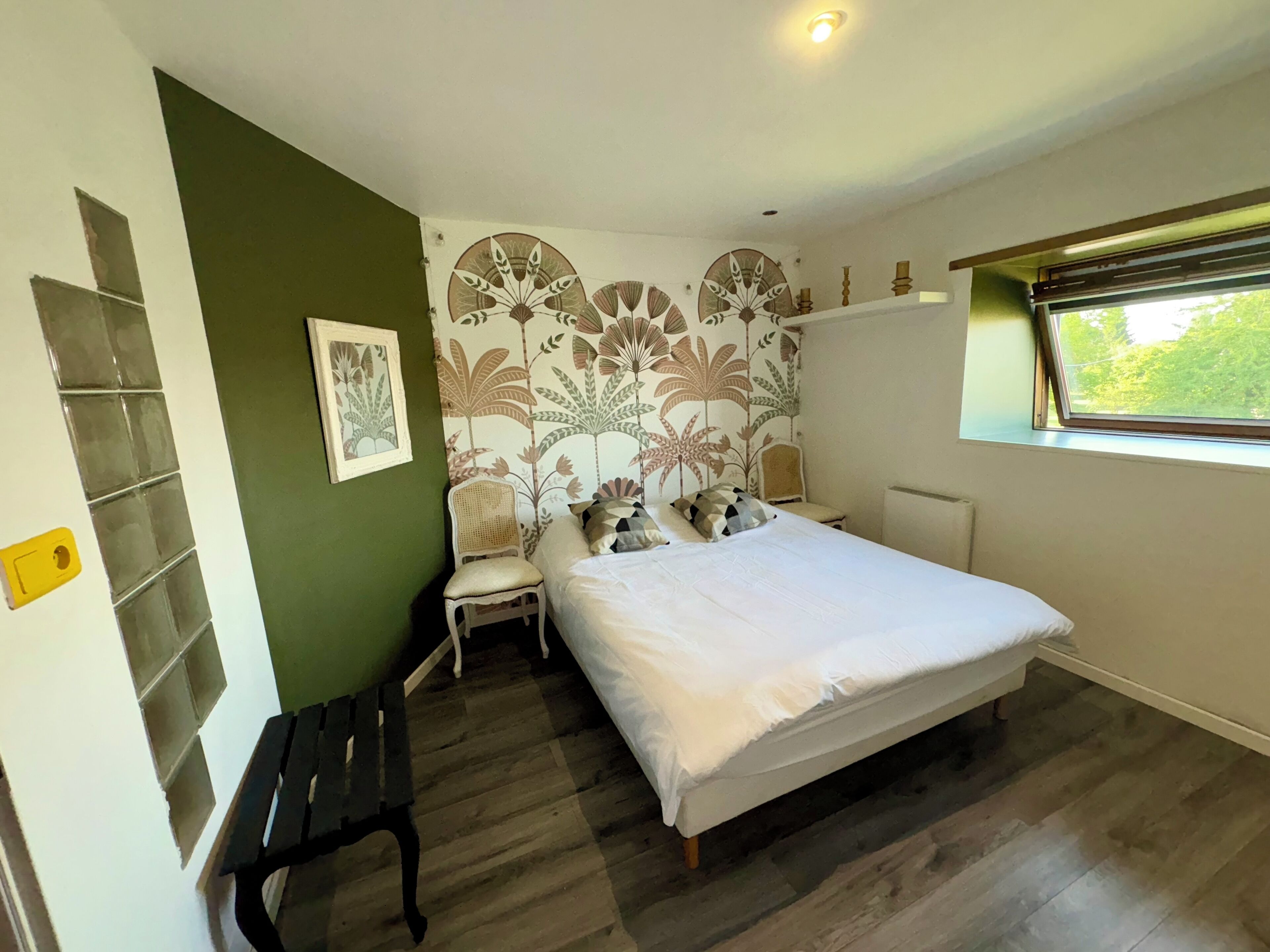 Une chambre à coucher comprenant un lit, une chaise, un banc, une fenêtre et un mur orné d’un motif floral.